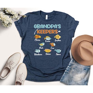 May include: A navy blue t-shirt with the text "Grandpa's Keepers" and a fishing rod with a line. There are six colorful fish with names written below them: Olivia, Kevin, Sophie, Paul, Eva, Madison, Adam, and Anna.