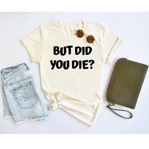 May include: A white t-shirt with the text "BUT DID YOU DIE?" printed in black. The shirt is laid flat on a white surface with a pair of sunglasses and a green clutch purse.