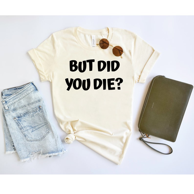 May include: A white t-shirt with the text "BUT DID YOU DIE?" printed in black. The shirt is laid flat on a white surface with a pair of sunglasses and a green clutch purse.