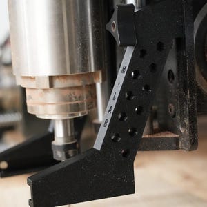 May include: A black metal router jig with a scale marked "M.L.A.E." and a knob for adjustment. The jig is attached to a router bit.