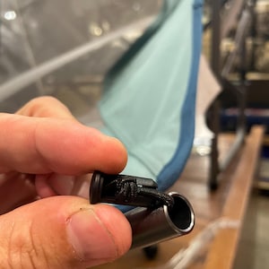May include: A black plastic cord lock attached to a thin black elastic string, which is inserted into the hollow end of a metallic gray tubular pole. The pole is connected to a light blue fabric panel that is part of a folding chair structure.