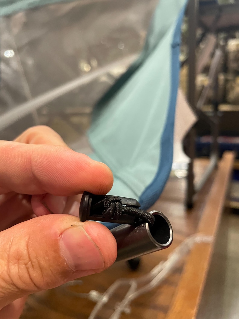 May include: A black plastic cord lock attached to a thin black elastic string, which is inserted into the hollow end of a metallic gray tubular pole. The pole is connected to a light blue fabric panel that is part of a folding chair structure.