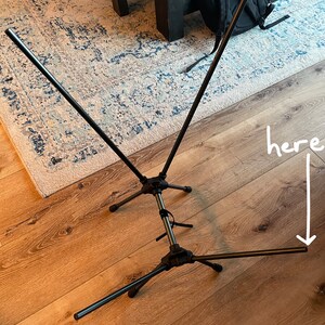 May include: A black collapsible metal stand featuring a central vertical pole with three tripod-style legs at the base and two angled support arms extending upward. The word here is written in white text with an arrow pointing to a leg tip.