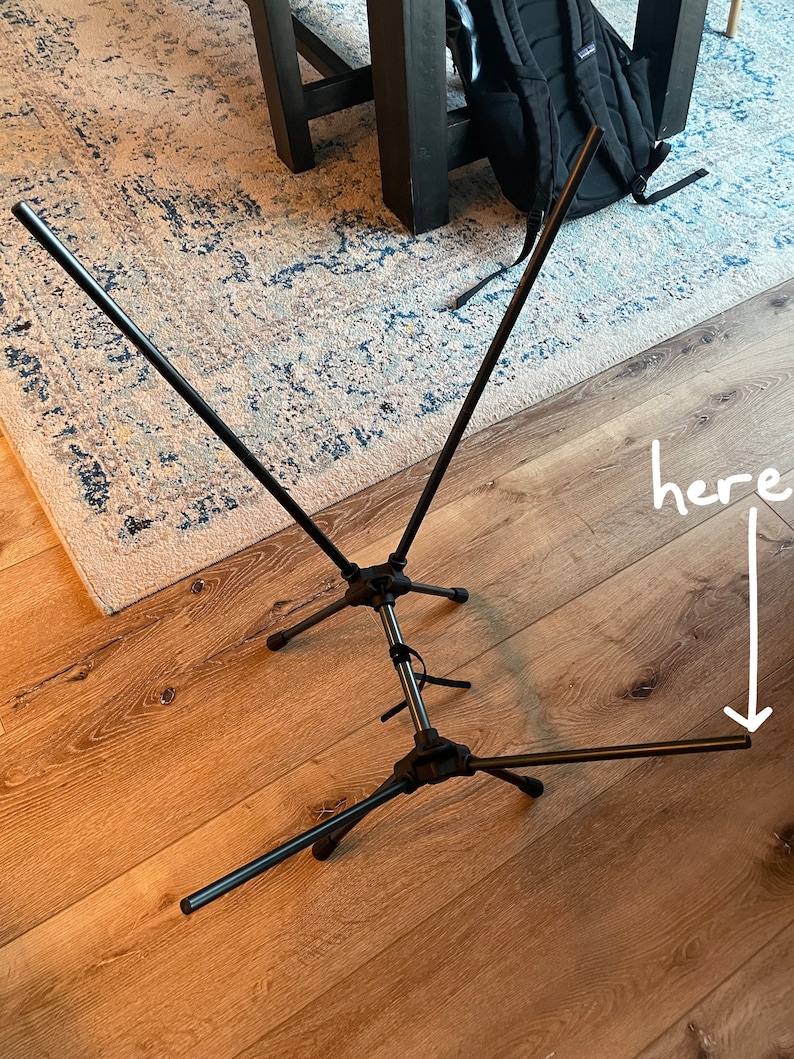 May include: A black collapsible metal stand featuring a central vertical pole with three tripod-style legs at the base and two angled support arms extending upward. The word here is written in white text with an arrow pointing to a leg tip.