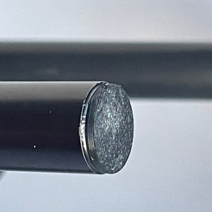 May include: A cylindrical black rod featuring a flat, circular end cap with a textured, metallic silver surface. The rod has a smooth, matte black finish and is positioned horizontally, showcasing the detailed, granular texture of the end piece.