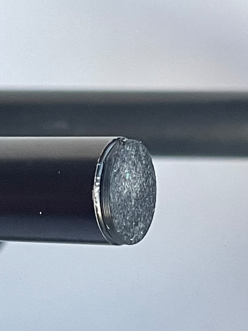 May include: A cylindrical black rod featuring a flat, circular end cap with a textured, metallic silver surface. The rod has a smooth, matte black finish and is positioned horizontally, showcasing the detailed, granular texture of the end piece.