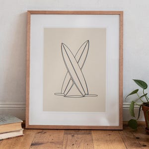 Crossed Surfboards Line print in a Red Oak frame with a white mat, standing on the floor, leaning back slightly against the wall in a living room floor lean.