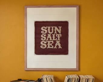 May include: A framed square textile art piece featuring a deep maroon felt background with the words SUN SALT SEA embroidered in cream-colored thread. The artwork is encased in a light-toned wooden frame with a wide off-white matting border.