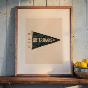 May include: A framed print featuring a dark green triangular pennant with the text OUTER BANKS and a small wave icon in white. The pennant has three small fabric tabs on the left side and is centered within a light-colored mat and a wooden frame.