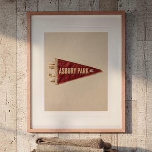 May include: A framed print featuring a red triangular pennant with the text ASBURY PARK and a small wave icon. The pennant has a textured, weathered appearance and is centered within a light-colored mat inside a thin, light-toned wooden frame.