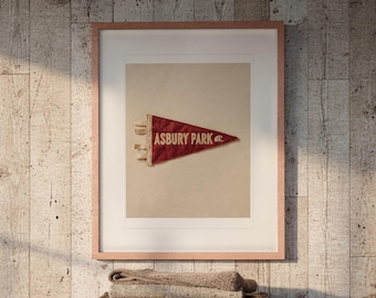 May include: A framed print featuring a red triangular pennant with the text ASBURY PARK and a small wave icon. The pennant has a textured, weathered appearance and is centered within a light-colored mat inside a thin, light-toned wooden frame.