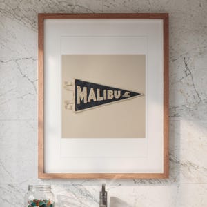 May include: A framed wall art piece featuring a triangular dark blue felt pennant with the word MALIBU printed in white capital letters and a small white wave graphic, displayed in a light brown wooden frame against a white background.