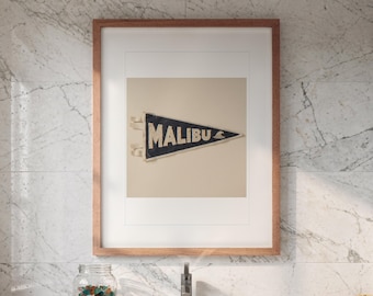May include: A framed wall art piece featuring a triangular dark blue felt pennant with the word MALIBU printed in white capital letters and a small white wave graphic, displayed in a light brown wooden frame against a white background.