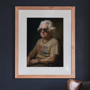 May include: A framed portrait of an older individual with white hair wearing dark sunglasses and a tan t-shirt with a graphic print. The artwork is encased in a light-toned wooden frame with a white mat, displayed against a dark wall.