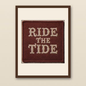 May include: A framed patch with the words "RIDE THE TIDE" embroidered in off-white thread on a maroon square. The patch is set against a cream background within a dark brown frame.