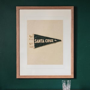 May include: A framed vintage-style dark green felt pennant featuring the text SANTA CRUZ in cream-colored lettering. The pennant is mounted on a light cream background within a light wood frame, displaying three small fabric ties on the left side.