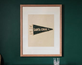 Pode incluir: Uma flâmula de feltro verde-escura de estilo vintage emoldurada, apresentando o texto SANTA CRUZ em letras cor de creme. A flâmula está montada sobre um fundo creme claro dentro de uma moldura de madeira clara, com três laços de tecido à esquerda.