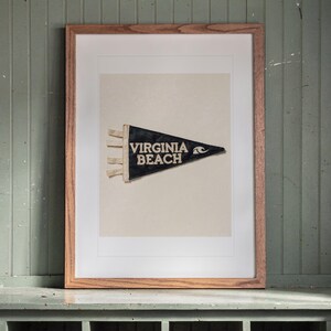 May include: A framed triangular felt pennant featuring the text VIRGINIA BEACH in white capital letters, accompanied by a white wave graphic. The pennant is dark blue with three white ties on the left side, displayed within a light wood frame.