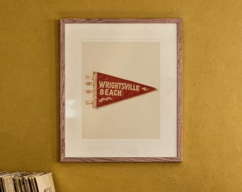 以下が含まれることがあります： WRIGHTSVILLE BEACHの白い文字と小さな波のグラフィックが描かれた、赤い三角形のフェルト製ペナント。明るい色のマットと木製のフレームに収められ、マスタードカラーの壁に飾られている。