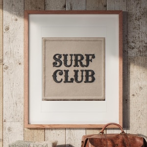 May include: A framed wall art piece featuring a square, textured beige fabric patch with the words SURF CLUB embroidered in bold, dark brown, decorative lettering. The artwork is enclosed in a simple, light-toned wooden frame.