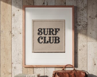Puede incluir: Una pieza de arte mural enmarcada que presenta un parche de tela beige texturizado y cuadrado con las palabras SURF CLUB bordadas en letras decorativas de color marrón oscuro. La obra está dentro de un marco de madera clara.