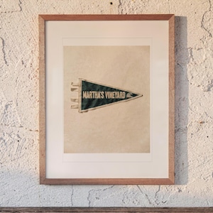 May include: A framed, dark green triangular felt pennant featuring the text MARTHA'S VINEYARD in white capital letters, accompanied by a small white wave graphic. The pennant is mounted on a light beige background within a light wood frame.