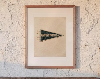 May include: A framed, dark green triangular felt pennant featuring the text MARTHA'S VINEYARD in white capital letters, accompanied by a small white wave graphic. The pennant is mounted on a light beige background within a light wood frame.