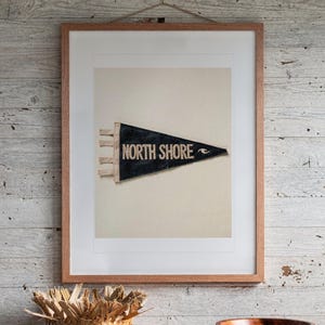 May include: A framed print featuring a black triangular pennant with the text NORTH SHORE in white capital letters and a small white wave icon. The pennant has three white ties on the left side and is centered within a light-colored mat and a wooden frame.