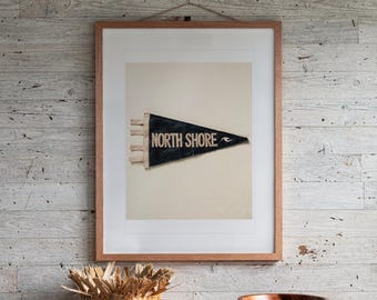 May include: A framed print featuring a black triangular pennant with the text NORTH SHORE in white capital letters and a small white wave icon. The pennant has three white ties on the left side and is centered within a light-colored mat and a wooden frame.