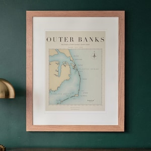 May include: A framed map print titled OUTER BANKS, featuring the coastline of North Carolina's barrier islands. The map displays Pamlico Sound and the Atlantic Ocean in light blue, with land in beige, housed in a light wood frame.