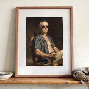 May include: A framed portrait print featuring a person wearing dark sunglasses and an unbuttoned short-sleeved patterned shirt, seated in a wooden chair. The artwork is displayed in a light brown wooden frame with a white mat border.