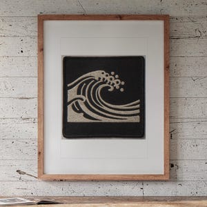 May include: A square embroidered patch featuring a stylised beige wave design on a solid black background, framed in a light brown wooden frame with a white mat, displayed as a piece of wall art.