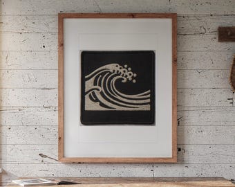 Puede incluir: Un parche bordado cuadrado que presenta un diseño de ola beige estilizado sobre un fondo negro sólido, enmarcado en un marco de madera marrón claro con un paspartú blanco, exhibido como arte de pared.
