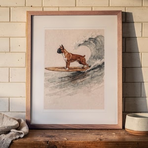 Puede incluir: Una lámina de acuarela enmarcada que muestra a un perro bóxer marrón y blanco de pie sobre una tabla de surf de madera montando una gran ola. La obra está en un marco de madera clara con paspartú blanco, mostrando una ilustración canina.