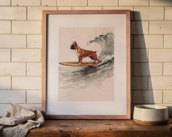 May include: A framed watercolour print featuring a brown and white Boxer dog standing on a wooden surfboard riding a large ocean wave. The artwork is enclosed in a light-toned wooden frame with a white mat, displaying a detailed canine illustration.