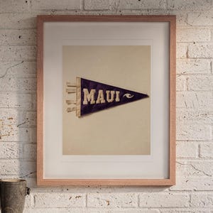 May include: A framed print featuring a dark purple felt pennant with the word MAUI in cream-colored block letters and a small wave graphic. The pennant has three cream-colored ties on the left side and is displayed in a light wood frame.
