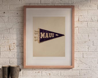 May include: A framed print featuring a dark purple felt pennant with the word MAUI in cream-colored block letters and a small wave graphic. The pennant has three cream-colored ties on the left side and is displayed in a light wood frame.
