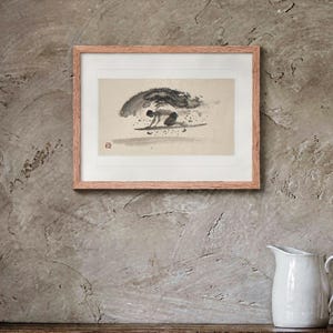 May include: A framed ink wash painting on paper depicting a person surfing on a wave. The artwork features a light beige background with a dark gray figure on a surfboard, a large cresting wave, and a small red square seal in the lower left corner.