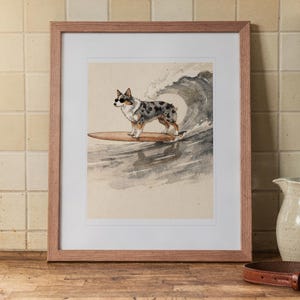 Puede incluir: Una lámina enmarcada con una ilustración en acuarela de un pastor australiano blue merle con gafas de sol sobre una tabla de surf surfeando una gran ola. La obra está en un marco de madera de color marrón claro con un paspartú blanco.