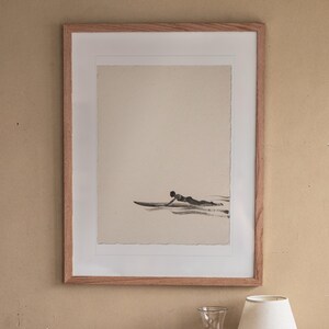 Paddling Surfer Ink Style print in a Red Oak frame with a white mat, hanging above the bedside table in a calm bedroom.