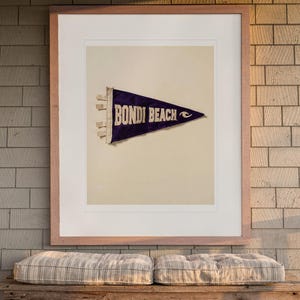 May include: A framed wall art print featuring a dark purple triangular felt pennant with the text BONDI BEACH in white capital letters and a small white wave graphic, displayed in a light wood frame against a brick wall.