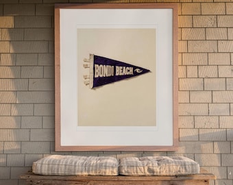 Puede incluir: Una impresión artística enmarcada que presenta un banderín triangular de fieltro de color morado oscuro con el texto BONDI BEACH en letras mayúsculas blancas y un pequeño gráfico de una ola blanca, en un marco de madera clara.