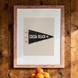 May include: A framed print featuring a dark blue triangular pennant with the text COCOA BEACH and a wave icon in cream lettering. The pennant has three cream-colored ties on the left side and is centered within a light wood frame.