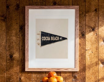 May include: A framed print featuring a dark blue triangular pennant with the text COCOA BEACH and a wave icon in cream lettering. The pennant has three cream-colored ties on the left side and is centered within a light wood frame.