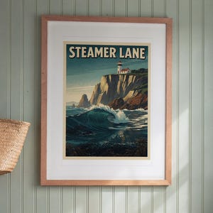 May include: A framed wall art print titled STEAMER LANE featuring a coastal scene with a lighthouse perched on a high cliff overlooking a large breaking ocean wave. The artwork uses a palette of deep blues, greens, and warm beige tones.
