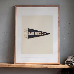 San Diego Surf Pennant print in a Red Oak frame with a white mat, leaning on the ledge against the wall in a hallway.
