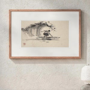 May include: A framed horizontal art print featuring a black ink wash illustration of a surfer riding a large wave. The artwork is set within a white mat and a light brown wooden frame, displaying a small red seal mark in the lower left corner.