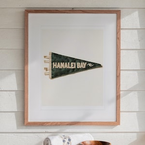 May include: A framed print featuring a dark green triangular pennant with the text HANALEI BAY and a small wave graphic in white lettering. The pennant has three white fabric ties on the left side and is displayed in a light wood frame.