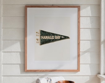 May include: A framed print featuring a dark green triangular pennant with the text HANALEI BAY and a small wave graphic in white lettering. The pennant has three white fabric ties on the left side and is displayed in a light wood frame.
