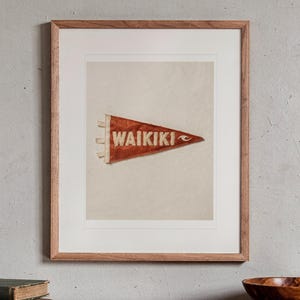 May include: A framed print featuring a triangular red felt pennant with the word WAIKIKI printed in white capital letters and a small white wave graphic on the right side. The pennant has three white fabric ties on the left edge.